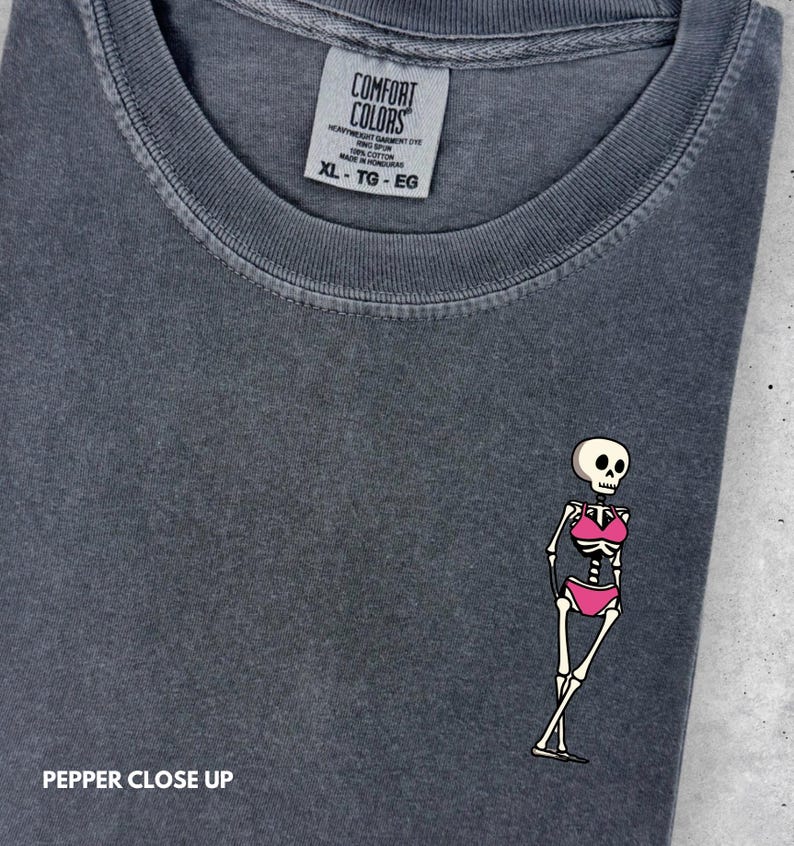 May include: A charcoal gray t-shirt with a crew neckline and a Comfort Colors brand label. The shirt features a small graphic of a skeleton wearing a pink bikini. The text PEPPER CLOSE UP is visible in the bottom left corner of the image.
