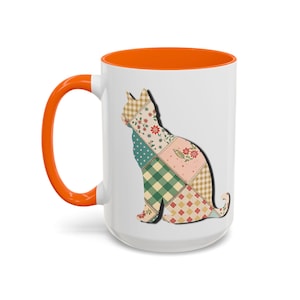 May include: White ceramic mug with an orange interior and handle. The mug features a patchwork cat silhouette in various patterns and colors. The cat design includes floral, polka dot, and plaid patterns.