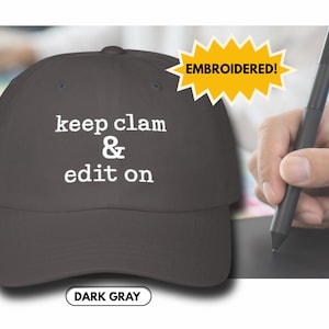 May include: Dark gray baseball cap with the embroidered text "keep clam & edit on" in white. The cap has a curved brim and a button on top. The text "EMBROIDERED!" is in a yellow starburst. The bottom of the cap says "DARK GRAY".