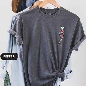 May include: A charcoal gray short-sleeve t-shirt featuring a small graphic on the left chest of a white skeleton wearing a bright pink bikini. The shirt has a crew neckline and a relaxed fit, with the word PEPPER printed in a black rectangular label.