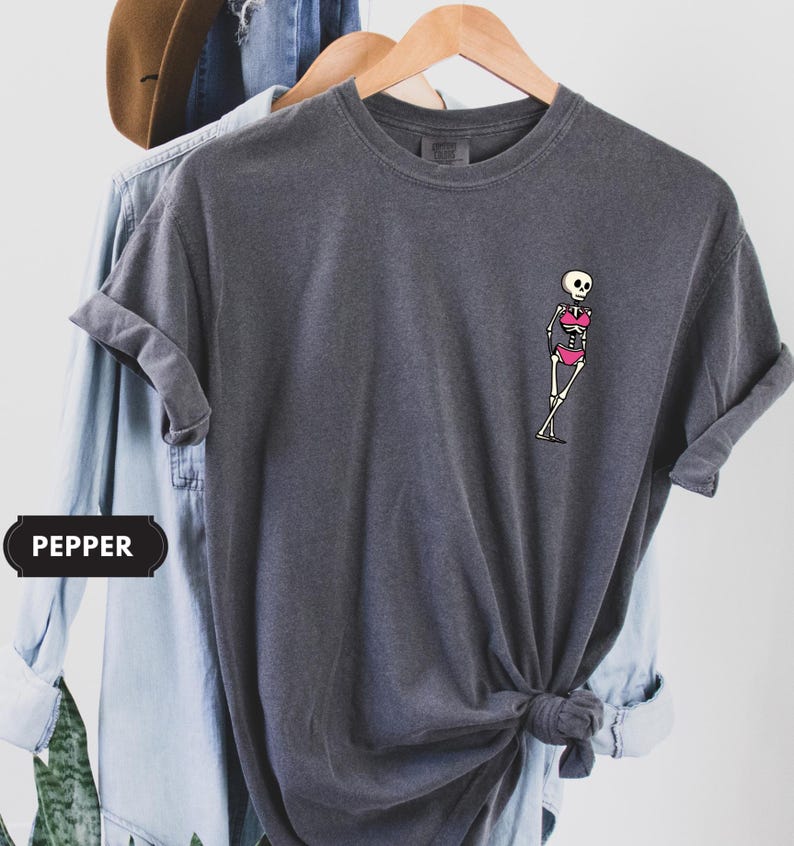 May include: A charcoal gray short-sleeve t-shirt featuring a small graphic on the left chest of a white skeleton wearing a bright pink bikini. The shirt has a crew neckline and a relaxed fit, with the word PEPPER printed in a black rectangular label.