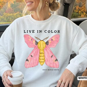 Puede incluir: Una sudadera blanca de cuello redondo con un gráfico de una polilla del arce rosada con alas de color rosa brillante y cuerpo amarillo. El texto "LIVE IN COLOR" está impreso en negro arriba y "ROSY MAPLE MOTH" está impreso en negro abajo.