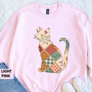 May include: Light pink sweatshirt featuring a patchwork cat design. The cat is composed of various patterned squares in shades of beige, red, green, and orange. The sweatshirt is a crew neck style, and the text "LIGHT PINK" is visible.