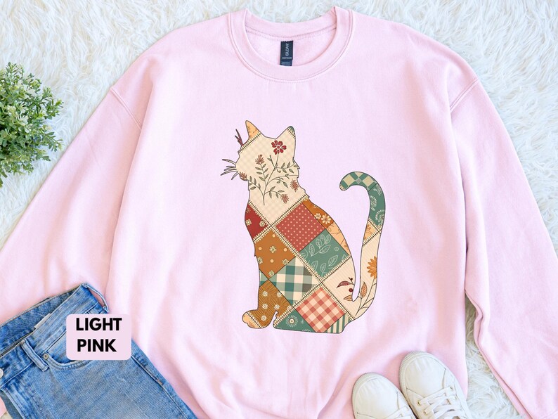 May include: Light pink sweatshirt featuring a patchwork cat design. The cat is composed of various patterned squares in shades of beige, red, green, and orange. The sweatshirt is a crew neck style, and the text "LIGHT PINK" is visible.