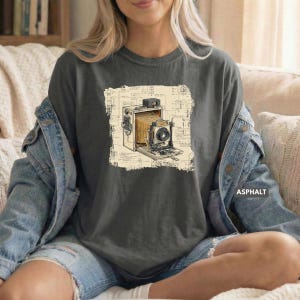 May include: A dark gray t-shirt featuring a graphic print of a vintage bellows camera centered on a background of technical blueprint sketches. The camera is depicted in shades of brown and black, showcasing its mechanical lens and folding structure.