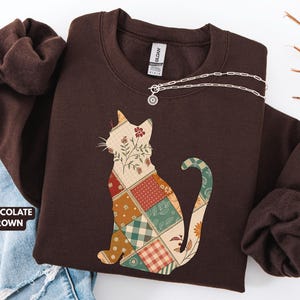 May include: Chocolate brown sweatshirt with a patchwork cat design. The cat silhouette is filled with various floral and plaid patterns. A silver necklace with a small pendant is draped across the sweatshirt.