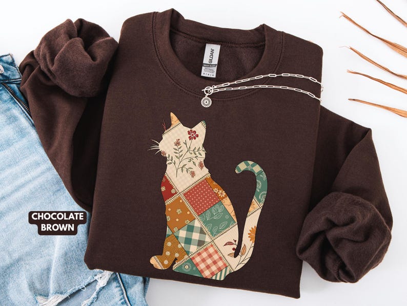 May include: Chocolate brown sweatshirt with a patchwork cat design. The cat silhouette is filled with various floral and plaid patterns. A silver necklace with a small pendant is draped across the sweatshirt.