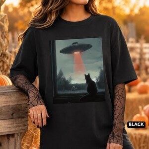 May include: A black short-sleeved t-shirt featuring a graphic print of a black cat sitting on a windowsill looking out at a flying saucer in the sky with a red light beam shining down. The shirt is worn over a long-sleeved black mesh top with a spiderweb pattern.