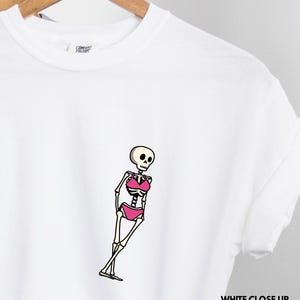 May include: A white t-shirt featuring a graphic of a skeleton wearing a pink bikini. The skeleton is depicted in a standing pose with its legs crossed. The shirt has a crew neckline and short sleeves, with a Comfort Colors brand label visible.