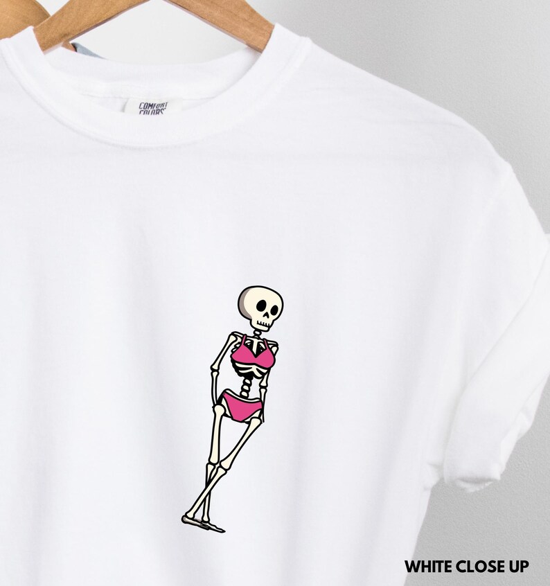 May include: A white t-shirt featuring a graphic of a skeleton wearing a pink bikini. The skeleton is depicted in a standing pose with its legs crossed. The shirt has a crew neckline and short sleeves, with a Comfort Colors brand label visible.
