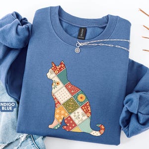 May include: Indigo blue sweatshirt with a patchwork cat design. The cat silhouette is composed of various floral and geometric patterns in red, yellow, green, and cream. A silver necklace with a pendant is on the neckline. The words "Indigo Blue" are visible.