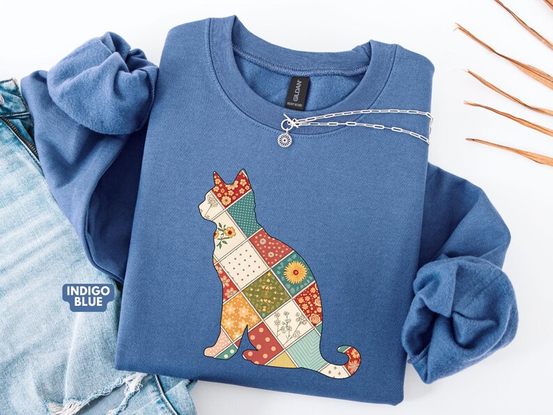 May include: Indigo blue sweatshirt with a patchwork cat design. The cat silhouette is composed of various floral and geometric patterns in red, yellow, green, and cream. A silver necklace with a pendant is on the neckline. The words "Indigo Blue" are visible.