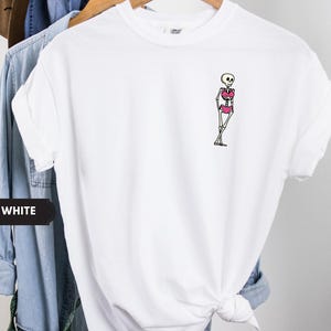 May include: A white short-sleeved t-shirt featuring a small graphic on the left chest area. The graphic depicts a cartoon skeleton wearing a bright pink bikini. The skeleton is standing with its legs crossed in a relaxed, leaning pose.