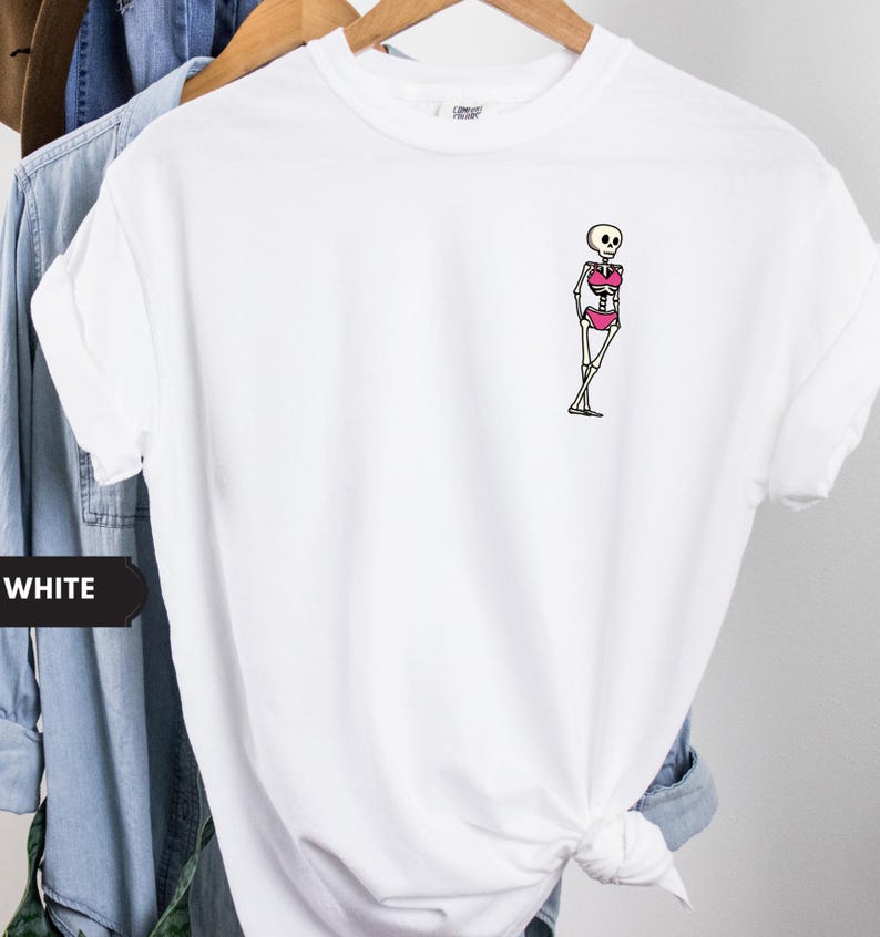 May include: A white short-sleeved t-shirt featuring a small graphic on the left chest area. The graphic depicts a cartoon skeleton wearing a bright pink bikini. The skeleton is standing with its legs crossed in a relaxed, leaning pose.