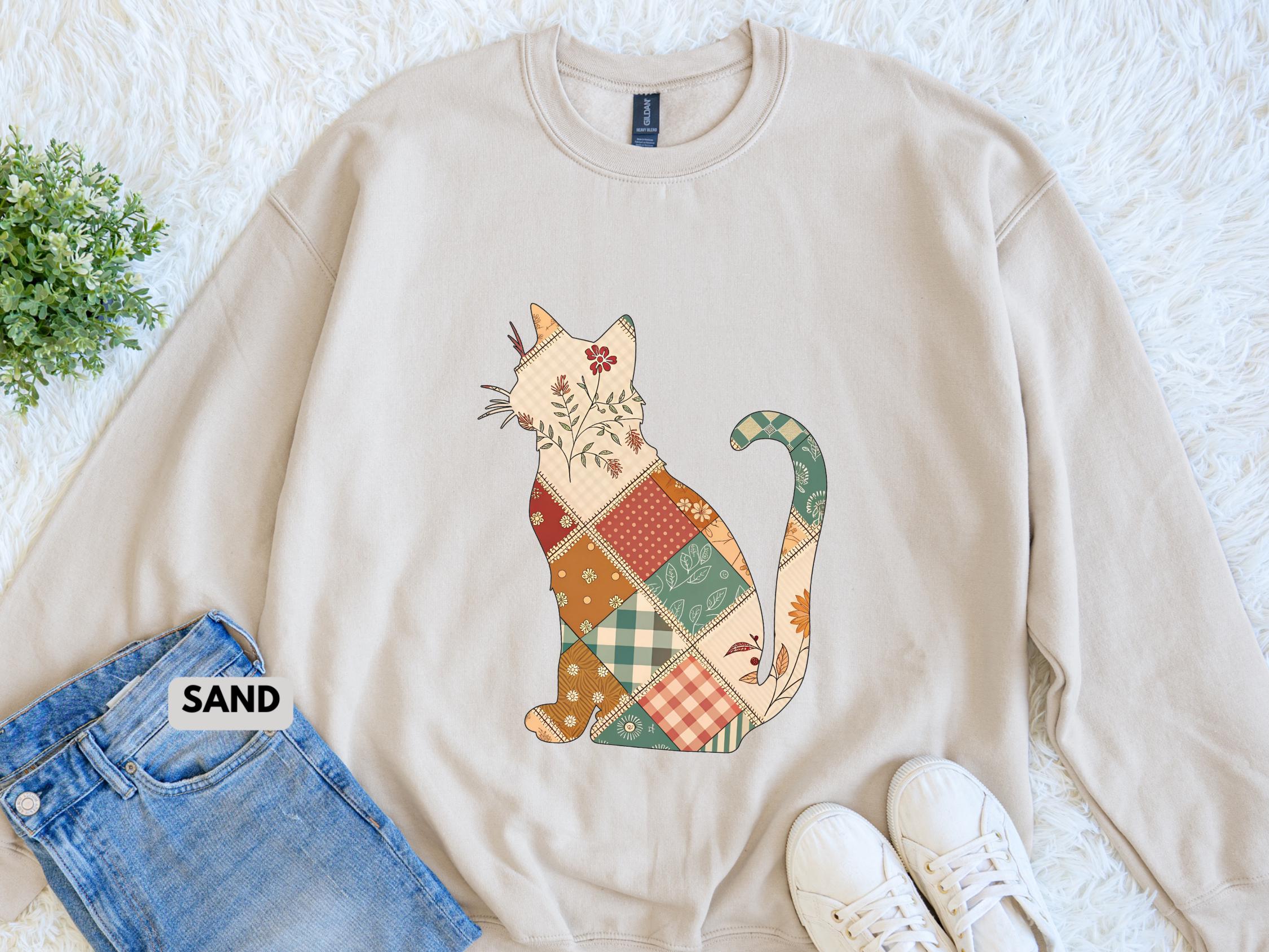 May include: Beige sweatshirt featuring a patchwork cat design. The cat is composed of various patterned squares in shades of red, green, and brown, with floral accents. The word "SAND" is visible on the bottom left.