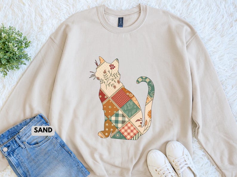 May include: Beige sweatshirt featuring a patchwork cat design. The cat is composed of various patterned squares in shades of red, green, and brown, with floral accents. The word "SAND" is visible on the bottom left.