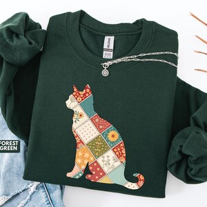 May include: Forest green sweatshirt featuring a patchwork cat design. The cat silhouette is filled with various floral and geometric patterns in red, yellow, and green. A silver necklace with a small pendant is draped across the neckline.