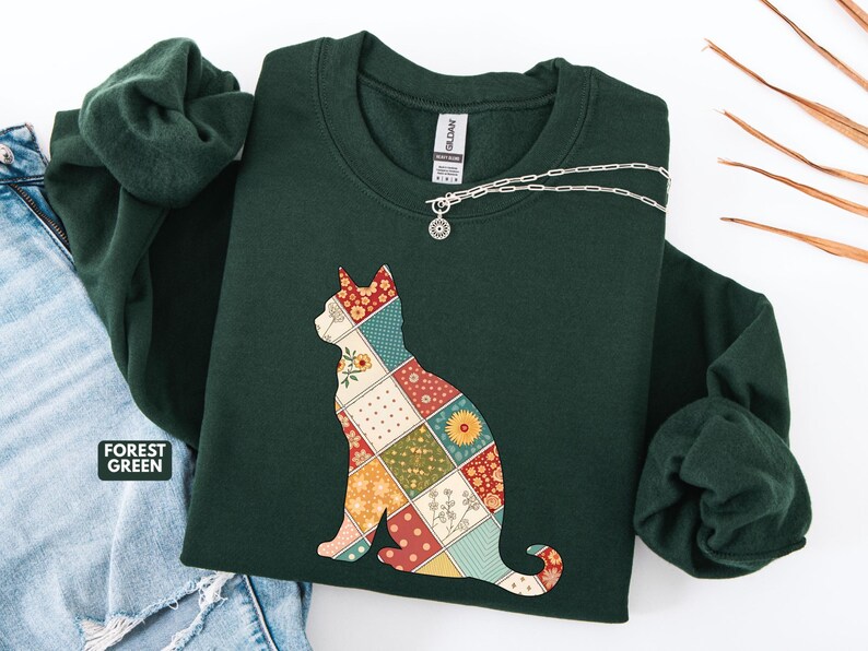May include: Forest green sweatshirt featuring a patchwork cat design. The cat silhouette is filled with various floral and geometric patterns in red, yellow, and green. A silver necklace with a small pendant is draped across the neckline.