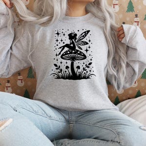 Op de afbeelding: Een grijs sweatshirt met een zwart silhouet van een fee die op een paddenstoel zit met sterren en bloemen eromheen.