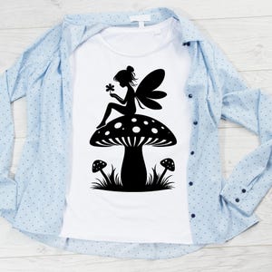 Op de afbeelding: Een wit T-shirt met een zwart silhouet van een fee die op een paddenstoel zit. De fee houdt een bloem vast en heeft vleugels. De paddenstoel heeft witte stippen en is omringd door gras.
