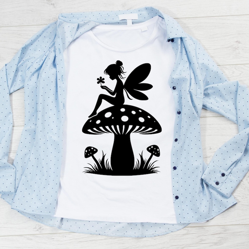 Op de afbeelding: Een wit T-shirt met een zwart silhouet van een fee die op een paddenstoel zit. De fee houdt een bloem vast en heeft vleugels. De paddenstoel heeft witte stippen en is omringd door gras.