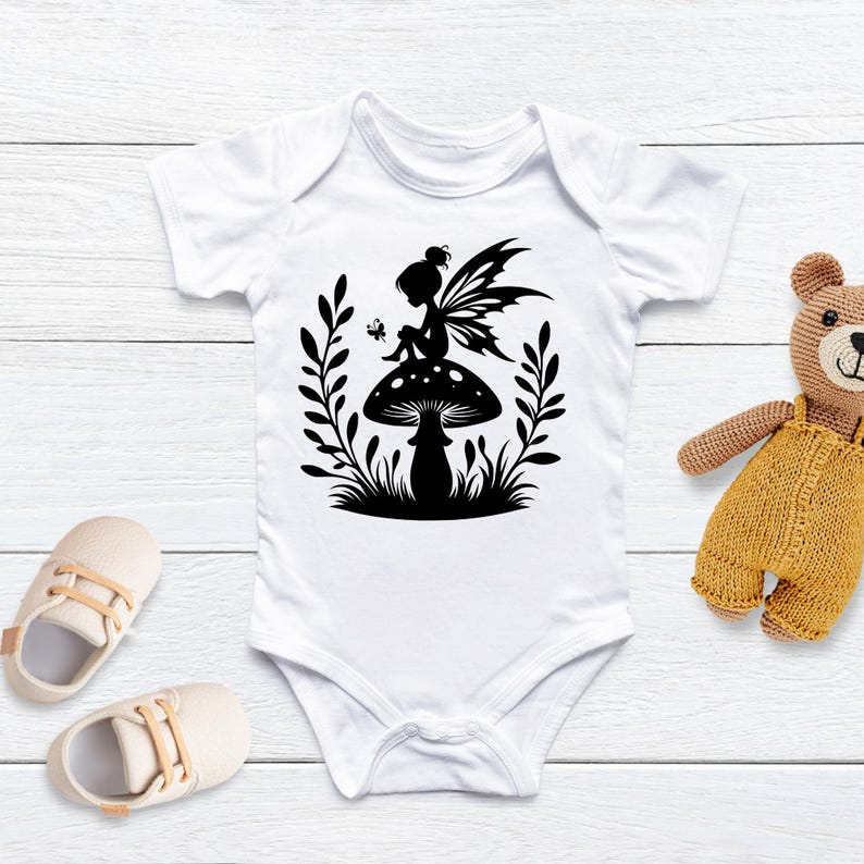 Op de afbeelding: Witte baby romper met een zwart silhouet van een fee die op een paddenstoel zit met uitgespreide vleugels. De fee is omringd door bladeren en een vlinder.