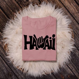 May include: A pink t-shirt with a black graphic of the word "HAWAII" with a hand gesture in the "A".