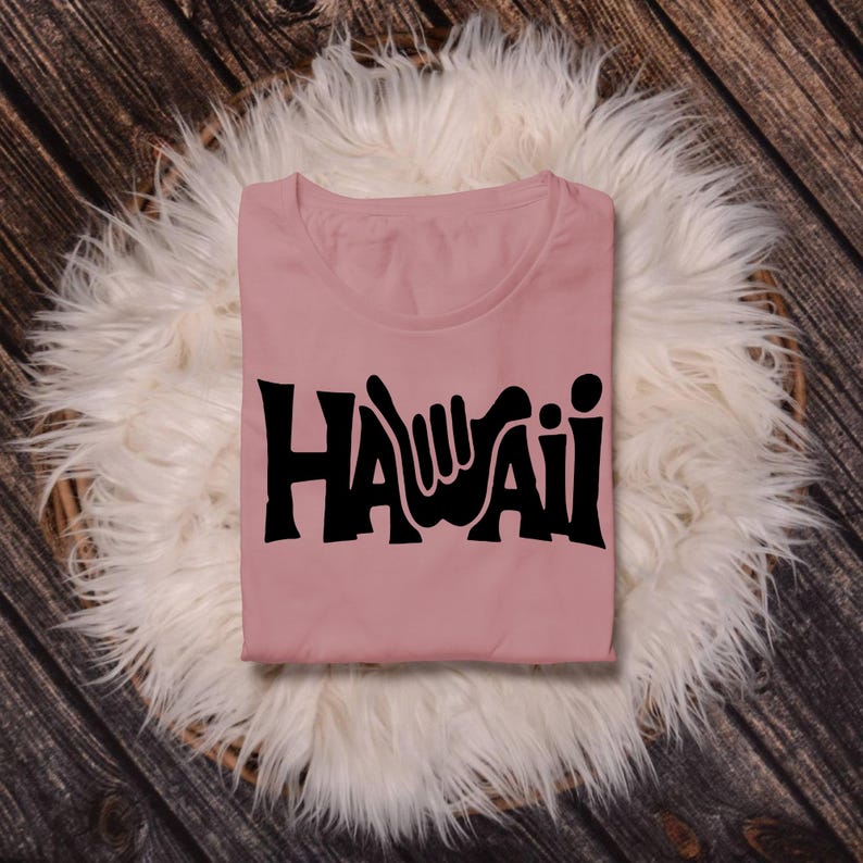 May include: A pink t-shirt with a black graphic of the word "HAWAII" with a hand gesture in the "A".