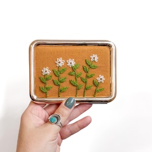 May include: A rectangular metal case with a polished silver finish featuring an embroidered fabric insert. The fabric is a mustard yellow color and displays five white daisy-like flowers with green stems and leaves stitched onto the surface.