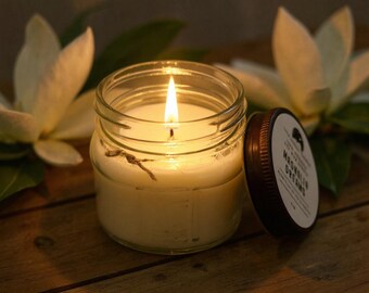 May include: A lit candle in a clear glass jar with a brown lid. The candle is surrounded by white flowers and green leaves. The lid has the words "Magnolia Dreams" printed on it.