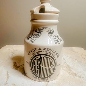 Puede incluir: Bote de mostaza de cerámica color crema con tapa. El bote presenta texto e ilustraciones en negro, incluyendo las palabras "FORTE AU VIN BLANC" y "CE POT de MOUTARDE". El bote tiene un asa en la tapa y un logotipo circular con texto.