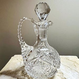 May include: A clear glass decanter with a faceted stopper and a cut glass design. The decanter has a handle and a round body.