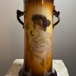 May include: A tall, antique vase with a brown and gold color scheme. The vase features a portrait of a woman in profile, adorned with jewelry. It has two ornate handles and a flared base, showcasing a vintage aesthetic.