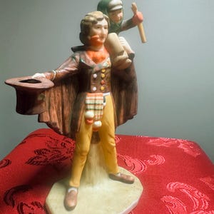 Puede incluir: Una figurita de porcelana de un hombre con un abrigo marrón y pantalones amarillos que sostiene un sombrero y lleva a un niño en sus hombros. El niño lleva un sombrero verde y sostiene un palo.