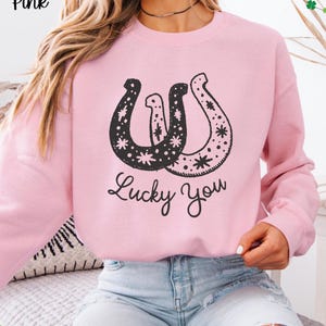 May include: Light pink sweatshirt with a black horseshoe design and the text "Lucky You". The horseshoe design is decorated with stars and dots. The words "Light Pink" are in the upper left corner.