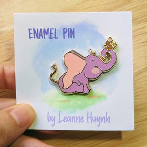 Puede incluir: Un pin de esmalte morado con un elefante de dibujos animados con orejas rosas y contorno dorado, sosteniendo un pequeño ratón. El pin está sobre una tarjeta blanca con el texto "ENAMEL PIN" y "by Leanne Huynh".