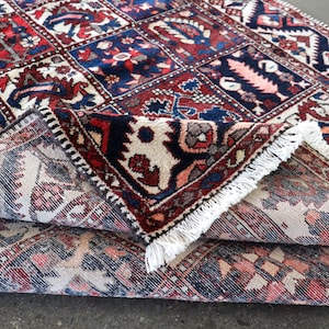 a close-up view of a colorful, intricately patterned rug with a mix of red, blue, and white colors. The rug has a fringe at the edges, adding to its decorative appearance. The rug appears to be folded or rolled up, revealing its detailed design.