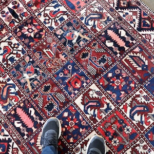 a colorful, intricately patterned rug with various designs and motifs, including floral and geometric patterns, arranged in a grid-like pattern. A pair of feet is visible standing on the rug, providing a sense of scale and perspective.