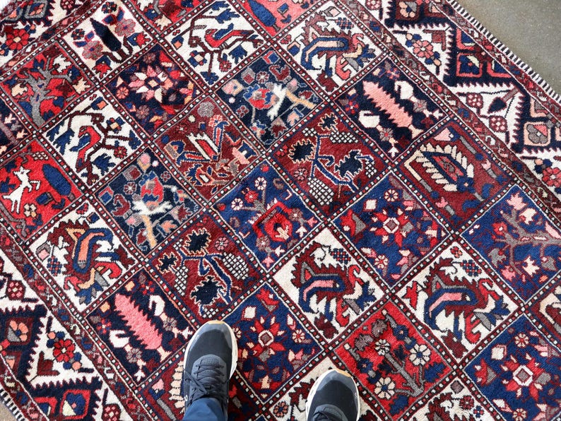 a colorful, intricately patterned rug with various designs and motifs, including floral and geometric patterns, arranged in a grid-like pattern. A pair of feet is visible standing on the rug, providing a sense of scale and perspective.