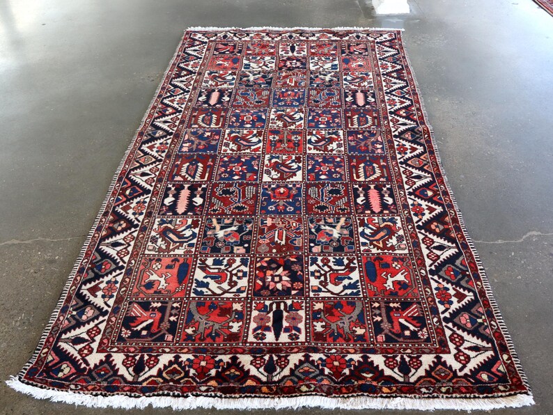a large, intricately designed rug with a vibrant pattern of red, blue, and white colors. The rug is placed on a concrete floor, and the background is blurred, drawing attention to the rug itself.