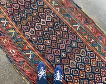 May include: A rectangular rug with a geometric pattern in shades of navy blue, red, orange, and teal. The rug features a central design of repeating octagons and a border with intricate patterns. The rug is placed on a concrete surface.
