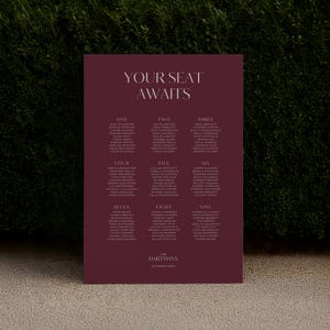 Op de afbeelding: Een bordeauxrode huwelijkszittingstabel met de tekst "YOUR SEAT AWAITS" bovenaan. De tabel is verdeeld in negen secties, elk met een nummer en een lijst met namen. De onderkant van de tabel luidt "The Hartmans 22 March 2025".