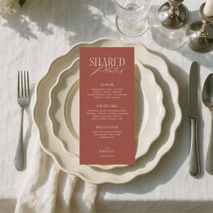 May include: A wedding menu card with the text "SHARED Plates" in a decorative font. The menu is printed on a rectangular, terracotta-colored card, placed on a white plate with a scalloped edge. Silverware and a water glass are also visible.