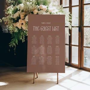 May include: A brown wedding guest list sign with the text "TAKE A SEAT" and "THE GUEST LIST" in a decorative font. The sign is supported by a wooden easel and is surrounded by floral arrangements.