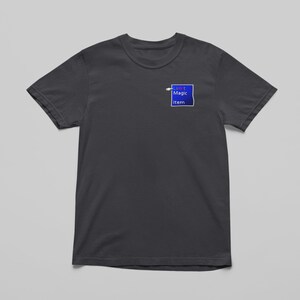 May include: Dark gray t-shirt with a crew neck. A small blue square with the text "My Magic Item" is printed on the upper left chest area. The shirt is made of a soft material and is displayed on a light gray background.