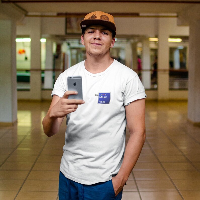 May include: A white t-shirt with a blue square patch that reads "Can't Magic Item". The person is holding a silver smartphone and wearing a brown baseball cap.