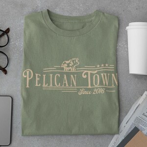 Op de afbeelding: Saliegroen T-shirt met de woorden "Pelican Town" in een sierlijke lettertype, een pixel koeienafbeelding en de tekst "Since 2016". Het shirt is opgevouwen op een grijze ondergrond.