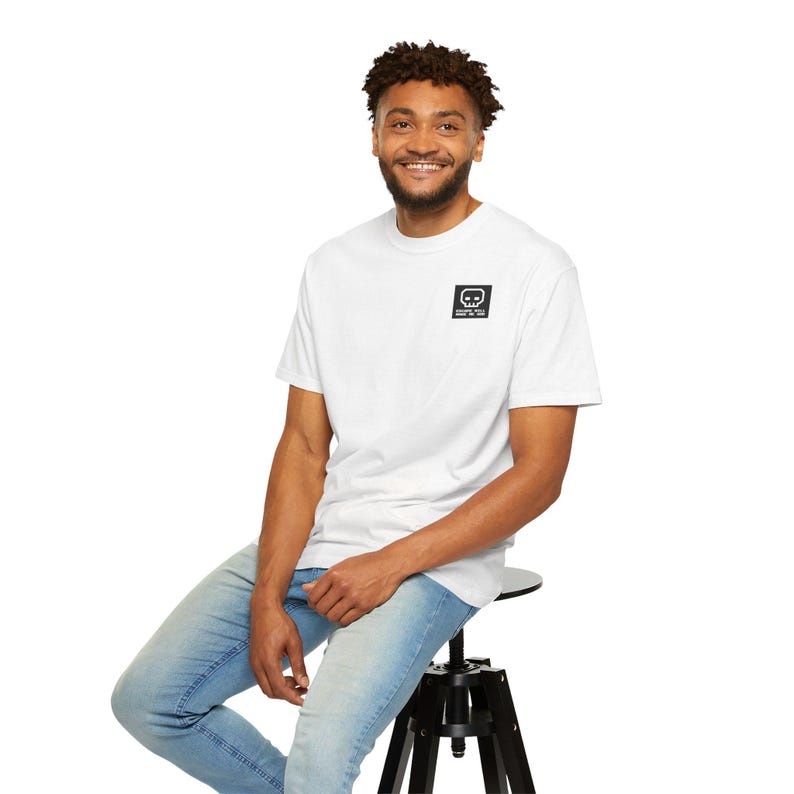 May include: White t-shirt with a small black square graphic on the chest. The graphic features a skull design and text. The person is wearing light blue jeans and is sitting on a black stool.