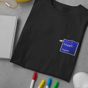 Puede incluir: Camiseta negra con un pequeño gráfico cuadrado en el pecho. El gráfico muestra las palabras "Limit Magic Item" en texto blanco sobre un fondo azul. La camiseta está sobre una superficie gris con un cuaderno, bolígrafos y un zapato.