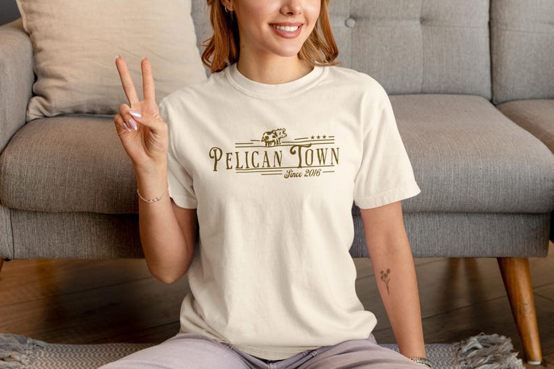 Op de afbeelding: Cr&egrave;mekleurig T-shirt met de tekst "Pelican Town Since 2016" in een bruin, vintage lettertype. Het ontwerp bevat een koeienafbeelding. Het shirt met korte mouwen wordt door een persoon gedragen.